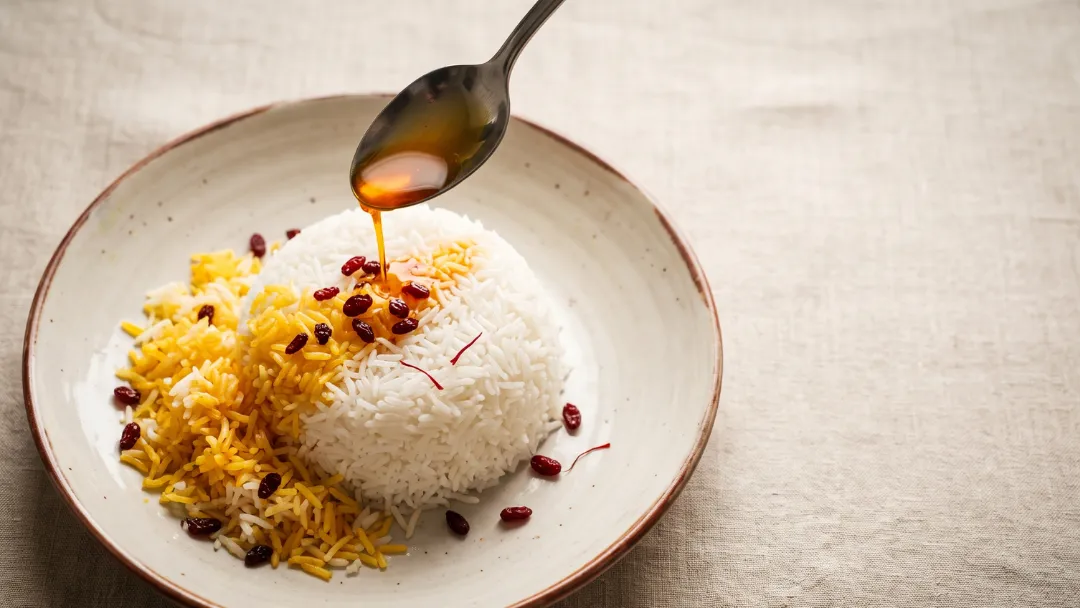 Réussir un riz au safran : eau de safran dorée versée à la cuillère sur un dôme de riz basmati blanc, un riz safrané doré à côté, parsemé de baies d'épine-vinette rouges et de filaments de safran, sur une assiette rustique.
