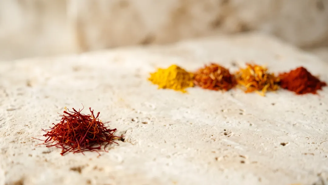 Substituts du safran : un tas de filaments de safran à l'écart, face à une rangée d'épices dorées (curcuma, carthame, fleur de souci, paprika) sur une pierre claire.